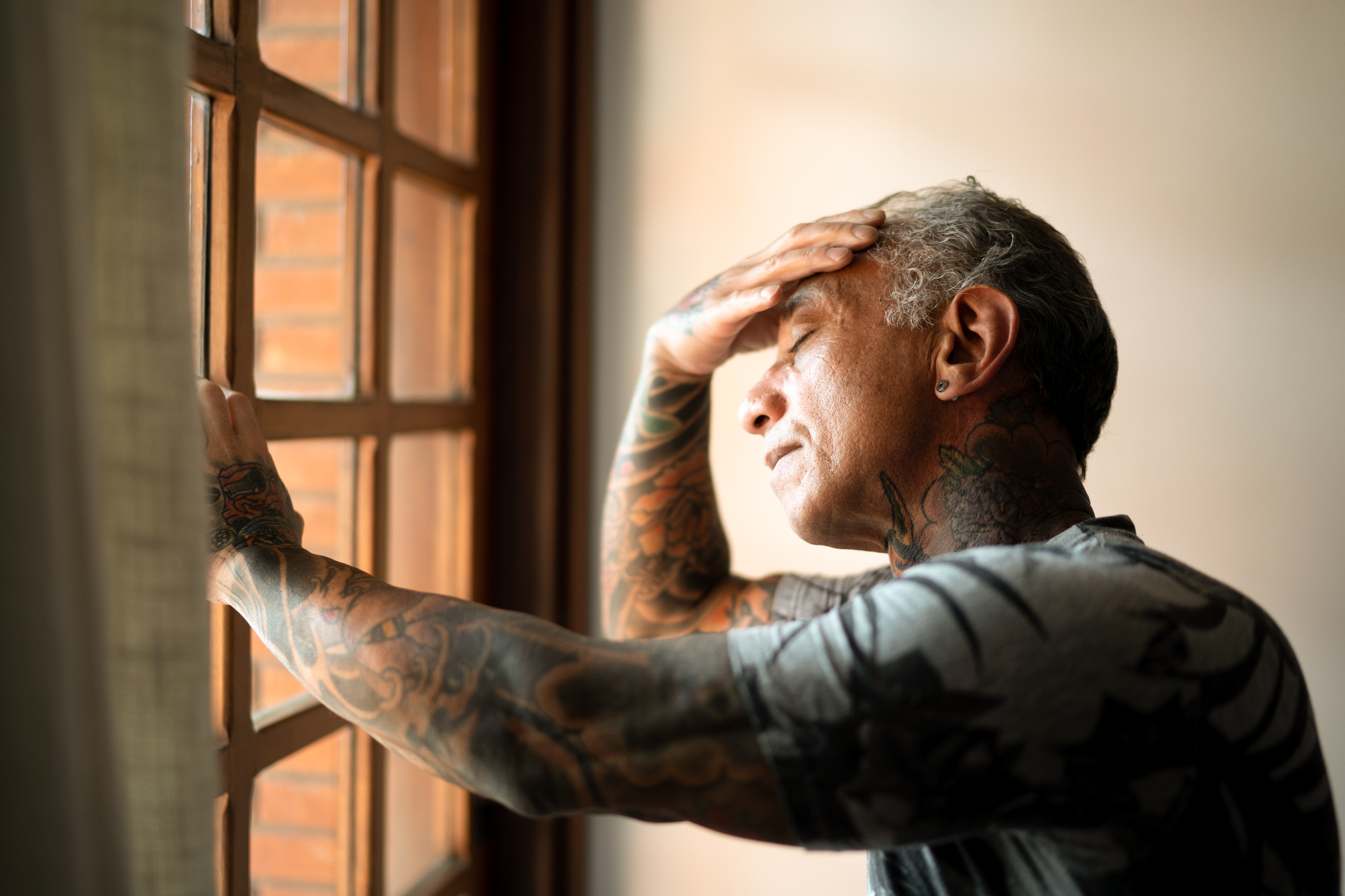 A person with dark skin tone supporting themselves against a window frame, one hand on their forehead, with eyes closed. 